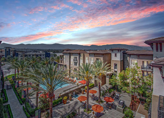 A serene Artisan at Main Street Metro apartment complex with palm trees, a pool, and orange umbrellas on a patio. The sky is vibrant with pink and blue hues, creating a tranquil mood.