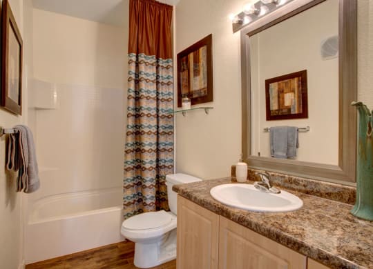 Modern bathroom here at Aspire at Pinnacle Peak Apartments with a white bathtub and a patterned curtain. A marble countertop has a sink, framed mirror, and decor. Neutral tones create a cozy feel.