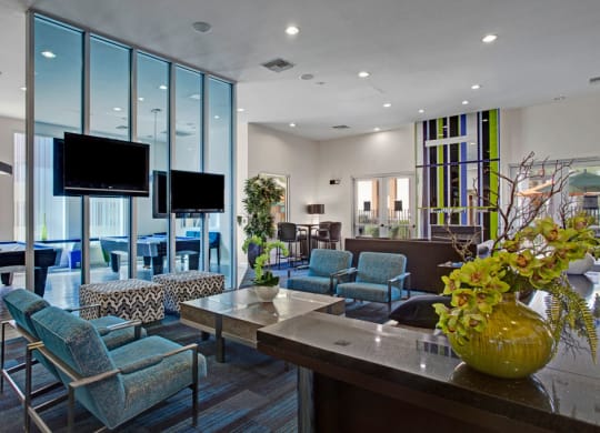 Modern lounge area here at Aspire at Pinnacle Peak Apartments with blue chairs, patterned ottomans, glass divider, and large TVs. Scenic decor includes green plants and a bright, spacious interior.