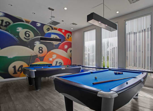 Modern billiards room here at Aspire at Pinnacle Peak Apartments with two blue pool tables, cue sticks, and balls. Features a colorful mural of large billiard balls and tall windows.