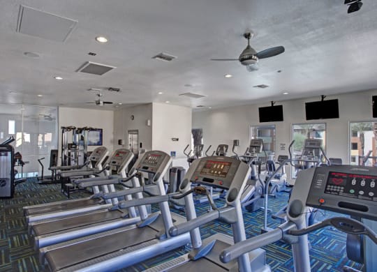 A modern gym here at Aspire at Pinnacle Peak Apartments with a row of treadmills and elliptical machines on a patterned carpet. Large windows and wall-mounted TVs create an open, bright atmosphere.