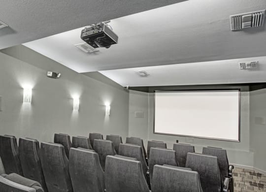 Small, dimly lit home theater here at Aspire at Pinnacle Peak Apartments with gray seats, a blank projector screen, and soft wall lighting, creating a cozy and intimate atmosphere.