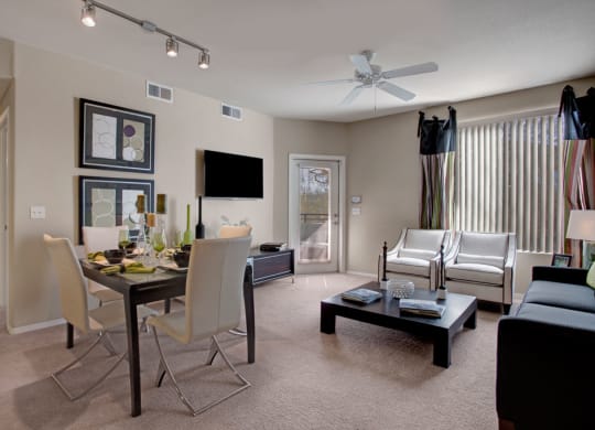 Modern living and dining space here at Aspire at Pinnacle Peak Apartments with neutral tones. A glass dining table with white chairs, abstract art, a wall-mounted TV, and cozy seating with natural light.