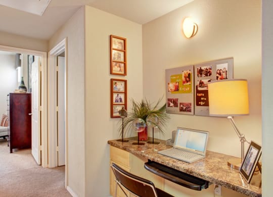 A cozy home office space here at Aspire at Pinnacle Peak Apartments with a marble desk, open laptop, modern chair, and a warm lamp. Family photos adorn the wall, creating a welcoming ambiance.