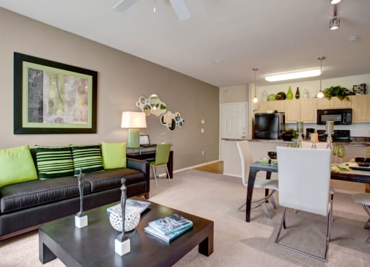 Modern living room here at Aspire at Pinnacle Peak Apartments with black sofa and green cushions, abstract art, and a sleek kitchen. Warm lighting and neutral tones create a cozy ambiance.