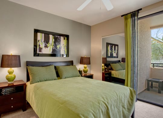 Modern bedroom here at Aspire at Pinnacle Peak Apartments with green-themed decor. Features include a queen bed, abstract artwork, matching lamps, wooden side tables, and a glass door leading to a balcony.