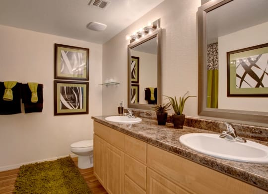 Modern bathroom here at Aspire at Pinnacle Peak Apartments with wood cabinets, marble countertop, dual sinks, large mirrors, abstract art, green towels, and a matching rug. Calm, clean ambiance.