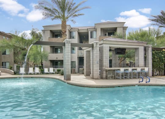 Luxurious pool area here at Aspire at Pinnacle Peak Apartments with clear blue water, lined with white lounge chairs. A modern cabana with a TV and bar seating creates a relaxed atmosphere.