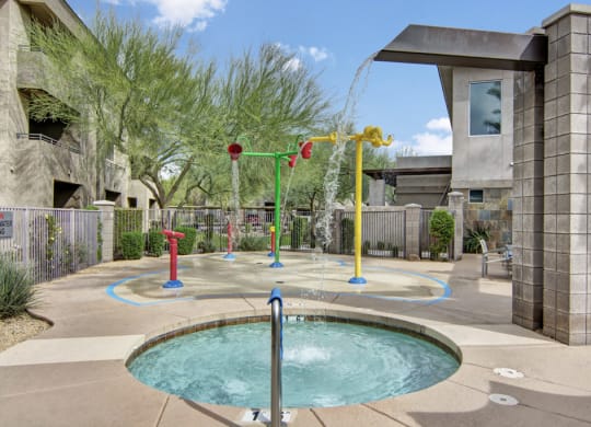 A vibrant children's splash pad here at Aspire at Pinnacle Peak Apartments with colorful water features and a small pool in a sunny outdoor setting. Surrounded by trees and concrete buildings.