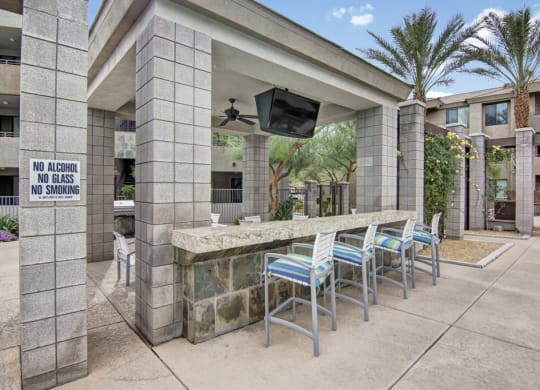 Outdoor lounge area here at Aspire at Pinnacle Peak Apartments with stone bar, colorful chairs, and TV under a shaded structure. Surrounded by palm trees, with a "No Alcohol, No Glass, No Smoking" sign.