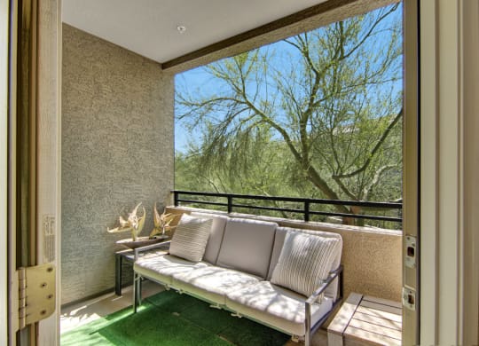 Cozy balcony with a cushioned sofa and striped pillows, overlooking a lush tree here at Aspire at Pinnacle Peak Apartments. Sunlight enhances the relaxed, natural ambiance.