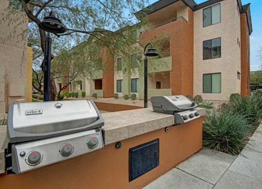 Two stainless steel barbecue grills here at Aspire at Pinnacle Peak Apartments are on a stone countertop in a modern apartment complex courtyard, surrounded by green plants and walking paths.