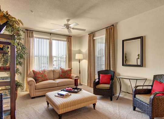 A warm and inviting living room here at Bela Rosa featuring a plush sofa with accent pillows, two woven armchairs with bold cushions, and a striped ottoman coffee table. Large windows with vertical blinds and curtains fill the space with natural light, while a ceiling fan, floor lamp, and wall mirror complete the comfortable and thoughtfully styled setting.