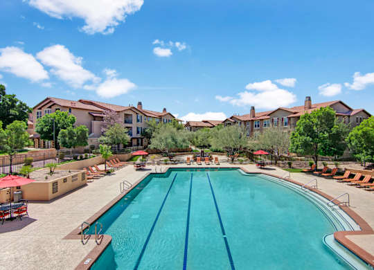 A resort-style swimming pool here at Bela Rosa featuring a spacious deck with multiple lounge chairs and red umbrellas for shaded relaxation. Clear blue water with lap lanes creates a refreshing focal point, while surrounding trees and warm-toned apartment buildings provide a private, inviting atmosphere perfect for enjoying sunny Arizona days.