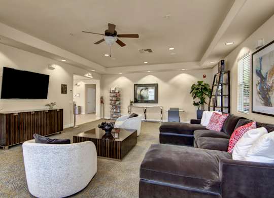 A welcoming resident lounge here at Bela Rosa featuring plush sectional seating with accent pillows, modern armchairs, and a sleek coffee table centered on patterned flooring. A large wall-mounted television, contemporary artwork, and bright windows create a comfortable space perfect for relaxing, socializing, or catching up with neighbors.