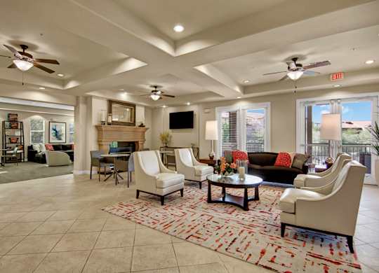 A spacious clubhouse lounge here at Bela Rosa featuring elegant armchairs and a plush sofa arranged around a round coffee table atop a patterned area rug. A cozy fireplace with mantel, wall-mounted television, and ceiling fans complement the open layout, while large windows and glass doors bring in natural light and create a bright, inviting gathering space.