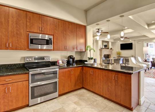 A modern kitchen here at Bela Rosa featuring rich wood cabinetry, sleek granite countertops, and stainless steel appliances including a built-in microwave and range. The open layout flows seamlessly into the clubhouse lounge beyond, while tile flooring, ample cabinet storage, and generous counter space create a functional and inviting space for everyday living and entertaining.