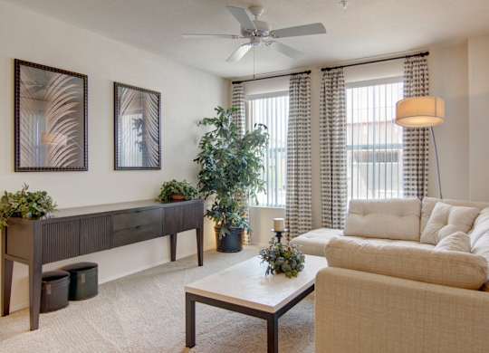 A bright and inviting living room here at Bela Rosa featuring plush neutral carpeting, a comfortable beige sofa, and a sleek coffee table centered beneath a ceiling fan. Large windows with patterned curtains bring in natural light, while framed wall art, a modern console table, and lush greenery add warmth and style to this cozy gathering space.