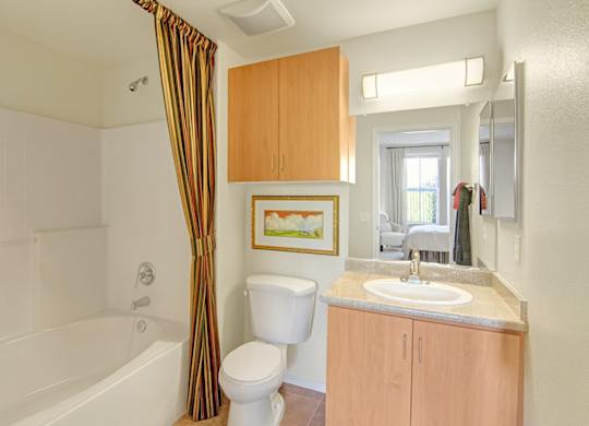 A clean and functional bathroom here at Bela Rosa featuring a white tub and shower combination with tiled surround and a colorful striped curtain. A light wood vanity with a smooth countertop and integrated sink offers storage below and above, while bright vanity lighting and a large mirror enhance the space, with a view into the adjoining bedroom beyond.