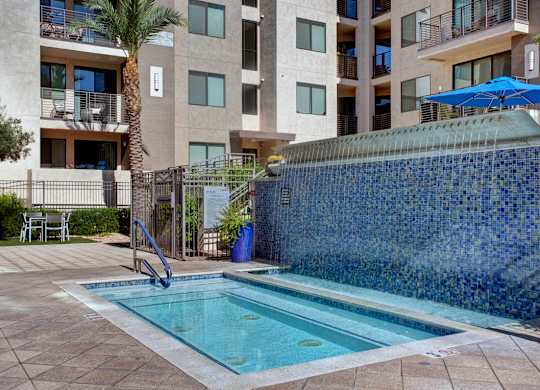 A contemporary spa area here at Escape featuring a tiled hot tub with built-in seating and handrails, complemented by a striking blue mosaic water feature wall, shaded lounge seating, and surrounding modern apartment homes with private balconies, creating a relaxing and upscale outdoor retreat space.