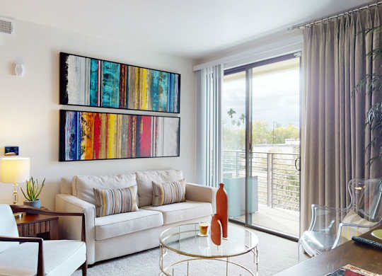 A bright contemporary living room here at Escape featuring a plush neutral sofa with patterned accent pillows, a round glass coffee table with decorative vases, and colorful abstract wall art, complemented by a cozy armchair and side table with lamp, while a sliding glass door with full-length curtains opens to a private balcony overlooking scenic views and palm trees.