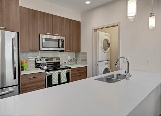 A contemporary open kitchen here at Escape showcasing rich wood-style cabinetry with sleek hardware, stainless-steel appliances including a built-in microwave and smooth-top range, a modern tile backsplash, and expansive quartz countertops with an undermount sink and gooseneck faucet, plus a convenient in-home washer and dryer neatly tucked into a nearby closet for added functionality.