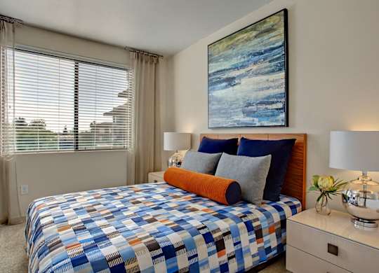 A stylish bedroom here at Escape featuring a warm wood headboard, layered blue and gray bedding with an accent bolster pillow, and matching nightstands topped with modern chrome lamps, while a large window with blinds and soft drapery fills the space with natural light and a bold abstract artwork adds a contemporary focal point above the bed.
