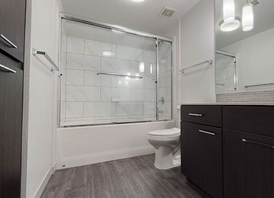A sleek modern bathroom here at Escape featuring a dark wood-style vanity with streamlined hardware, a wide mirror with contemporary lighting, and a spacious countertop with integrated sink, complemented by a tub and shower combination with glass enclosure and large tile surround, wood-style flooring, and clean neutral finishes for a polished, spa-inspired feel.