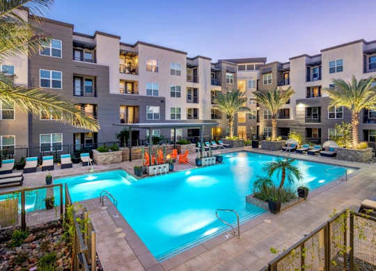 A resort-inspired pool courtyard here at The Flats at San Tan featuring a sparkling turquoise swimming pool with wide entry steps, surrounding lounge chairs with cushioned seating, illuminated palm trees, and a covered cabana with vibrant Adirondack chairs, all framed by contemporary apartment homes and warm evening lighting for a relaxing community retreat.