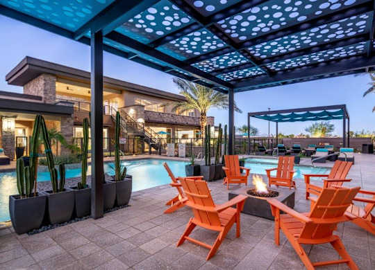 A stylish outdoor lounge area here at The Flats at San Tan featuring bright orange Adirondack chairs gathered around a modern fire pit beneath a patterned pergola, surrounded by a resort-style swimming pool, private cabanas, tall palm trees, and sleek contemporary clubhouse architecture illuminated by soft evening lighting.