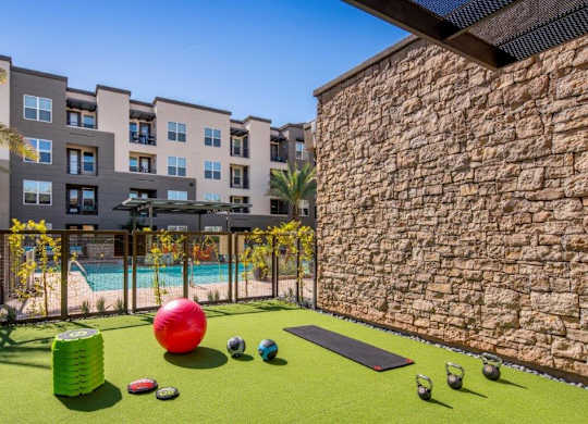 A vibrant outdoor fitness area here at The Flats at San Tan featuring green turf flooring with kettlebells, weighted balls, a yoga mat, and stacked step platforms, set beside a stone accent wall and overlooking the resort-style pool and modern apartment buildings with palm trees and private balconies.