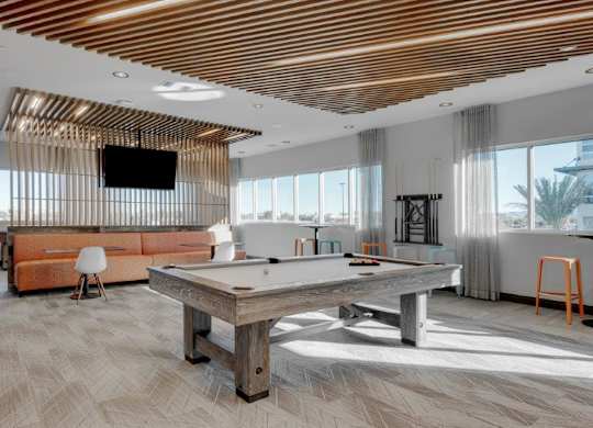 A stylish resident lounge here at The Flats at San Tan featuring a modern pool table on patterned tile flooring, wood slat ceiling accents, expansive windows with sheer curtains, and a cozy seating area with an orange sectional, mounted television, and high-top tables, creating an inviting space for games, relaxation, and social gatherings.