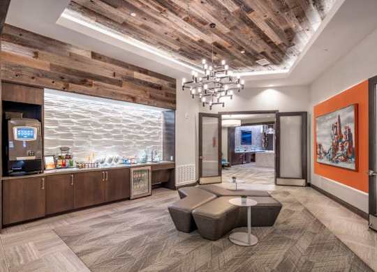 A contemporary resident lounge here at The Flats at San Tan showcasing a dramatic wood plank ceiling, modern chandelier lighting, and a built-in refreshment bar with cabinetry, countertop space, and beverage refrigerator, complemented by geometric lounge seating, glass double doors, and bold accent artwork that creates a welcoming space for gatherings and relaxation.