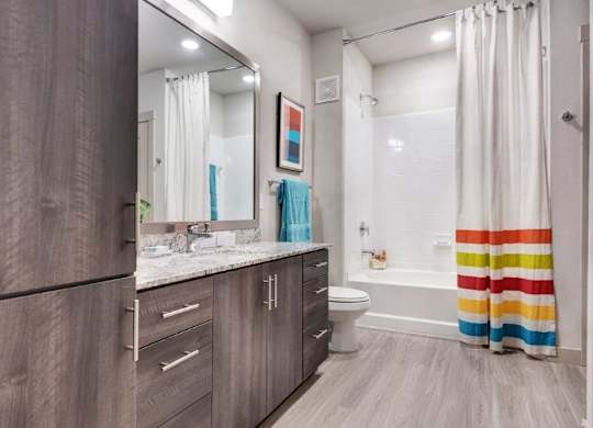 A bright and modern bathroom here at The Flats at San Tan showcasing wood-style cabinetry with sleek hardware, a spacious granite countertop with an undermount sink, and a large framed mirror that enhances the open feel, complemented by wood-look flooring, a white tub and shower combination, and a colorful striped shower curtain that adds a vibrant touch to the space.