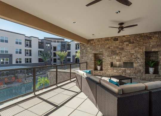 A spacious covered patio lounge here at The Flats at San Tan featuring woven sectional seating with plush cushions, a sleek outdoor coffee table, and a striking stone accent wall with a built-in fireplace, complemented by ceiling fans, recessed lighting, and a metal railing overlooking the sparkling pool and surrounding apartment homes for an inviting community gathering space.
