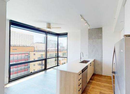 A sleek and contemporary kitchen here at JOY on 4th featuring light wood cabinetry, quartz-style countertops, and stainless-steel appliances, highlighted by a streamlined undermount sink and modern track lighting, while expansive floor-to-ceiling corner windows fill the space with natural light and showcase vibrant city views alongside warm wood-style flooring.