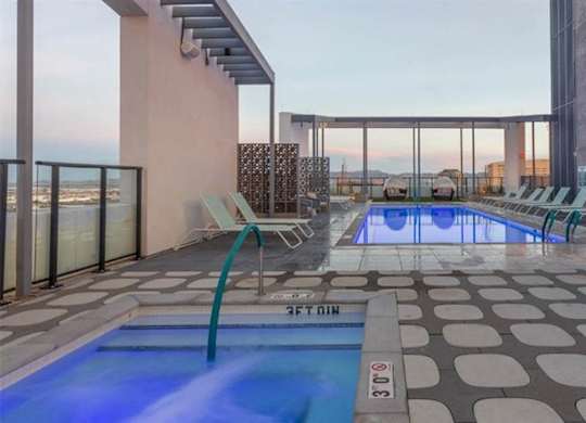 A modern rooftop pool and spa here at JOY on 4th featuring a glowing in-ground hot tub and a sleek lap pool lined with contemporary lounge chairs. Decorative privacy screens and pergola structures frame sweeping city and mountain views, while glass railings and patterned tile decking create a stylish, elevated retreat above downtown.