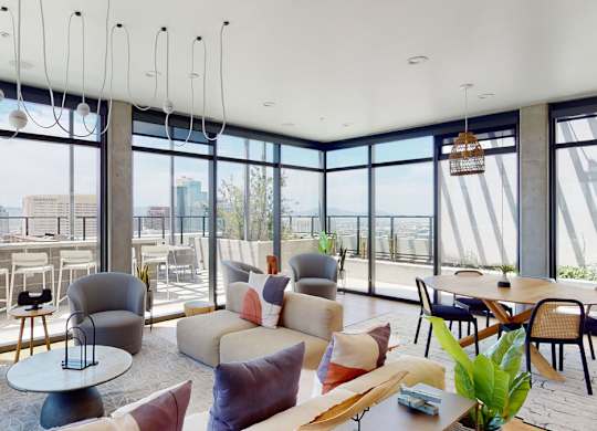 A panoramic rooftop lounge here at JOY on 4th showcasing floor-to-ceiling windows with sweeping skyline and mountain views, contemporary seating in soft neutral tones, and warm wood flooring. Stylish armchairs and a plush sofa surround modern tables, while a dining area and outdoor terrace seating create an open, light-filled space ideal for relaxing or gathering.