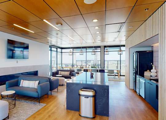 A sophisticated resident lounge here at JOY on 4th featuring warm wood ceilings with recessed lighting, expansive windows with skyline views, and sleek wood-style flooring throughout. Contemporary sofas and ottomans surround a modern linear fireplace, while a large quartz island, pendant lighting, and built-in kitchenette create an inviting space for gathering, working, or entertaining.