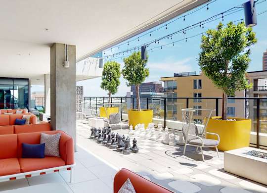 A vibrant rooftop terrace here at JOY on 4th featuring bold orange lounge seating, oversized yellow planters with lush trees, and string lighting overhead. A life-size chess set and café-style tables overlook surrounding city views, while sleek tile flooring and glass railings create a modern, open-air space perfect for relaxing or gathering with friends.