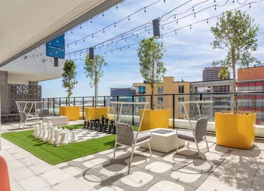 A lively rooftop terrace here at JOY on 4th featuring oversized yellow planters with tall trees, string lights overhead, and sweeping city views beyond sleek glass railings. Café-style tables and chairs sit beside a life-size chess set on green turf, creating a modern, open-air retreat perfect for socializing, relaxing, and enjoying the skyline.