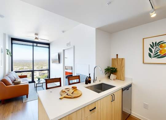 A bright and contemporary open kitchen and living space here at JOY on 4th showcasing light wood cabinetry, sleek quartz-style countertops, a stainless-steel undermount sink, and modern appliances, flowing seamlessly into a cozy living area with an orange sofa and floor-to-ceiling windows that frame expansive city views and fill the home with natural light.