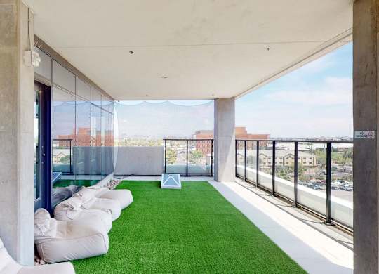A modern rooftop lounge here at JOY on 4th featuring lush green turf flooring, relaxed bean bag seating along a glass wall, and sleek concrete columns framing open skyline views. Glass railings overlook the city, while shaded overhead coverage creates a breezy, elevated retreat ideal for casual gatherings and outdoor relaxation.