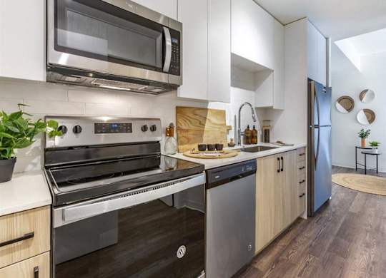 A sleek contemporary kitchen here at JOY on 4th featuring light wood cabinetry, white quartz countertops, and a modern tile backsplash. Stainless steel appliances, including a built-in microwave and full-size range, pair with a deep undermount sink and matte black hardware, while wood-style flooring flows into the open living area beyond.
