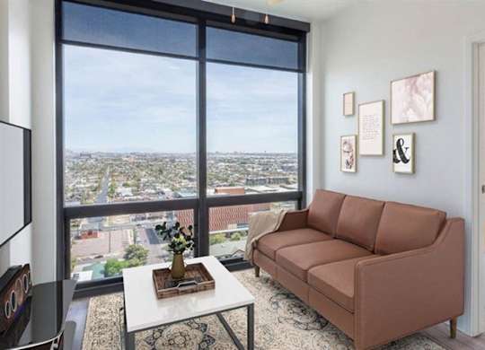 A bright modern living room here at JOY on 4th featuring a sleek brown sofa, patterned area rug, and contemporary wall art. Floor-to-ceiling windows showcase sweeping city and mountain views while filling the space with natural light, and an open doorway leads to a cozy bedroom with warm accents and expansive skyline scenery.