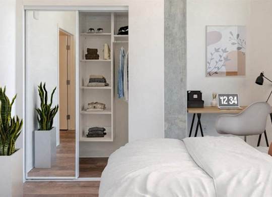 A bright modern bedroom here at JOY on 4th featuring soft neutral bedding, a warm accent pillow, and a sleek wall-mounted reading lamp above a compact desk workspace. Sliding mirrored closet doors reveal built-in shelving, while a tall potted plant and contemporary artwork add inviting style to this light-filled retreat.