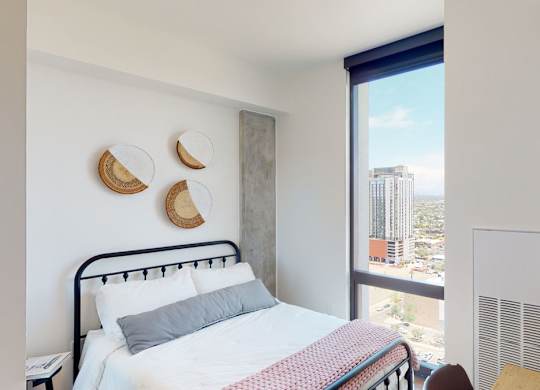 A bright and modern bedroom here at JOY on 4th featuring a sleek black metal bed frame with crisp white bedding and a soft accent throw, complemented by woven wall décor and a concrete accent column. Expansive floor-to-ceiling windows showcase sweeping city views while natural light enhances the clean lines, warm wood flooring, and contemporary finishes.