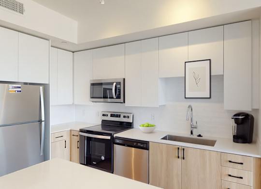 A modern and light-filled kitchen here at JOY on 4th showcasing sleek white upper cabinetry paired with warm wood lower cabinets and matte black hardware. Stainless steel appliances, smooth countertops, and a contemporary tile backsplash create a clean, streamlined look, while the deep undermount sink and generous prep space offer both style and everyday functionality.