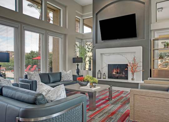 A welcoming clubhouse living room here at Mirador featuring soaring ceilings and expansive windows that fill the space with natural light. Contemporary leather seating surrounds a sleek coffee table and modern fireplace, while a mounted television and built-in shelving create a stylish, comfortable setting for relaxing or gathering with neighbors.