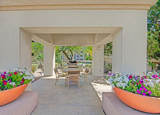 A shaded outdoor ramada here at Mirador featuring a covered concrete patio with a round dining table and chairs, surrounded by vibrant flower planters in large terracotta pots. A built-in grill and prep area sit just beyond the seating, creating a welcoming space for outdoor dining and relaxed gatherings among mature trees and landscaped grounds.